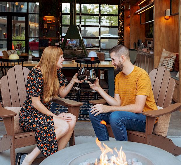 A couple toasting a glass of wine in brown adirondack chairs by a fire pit.